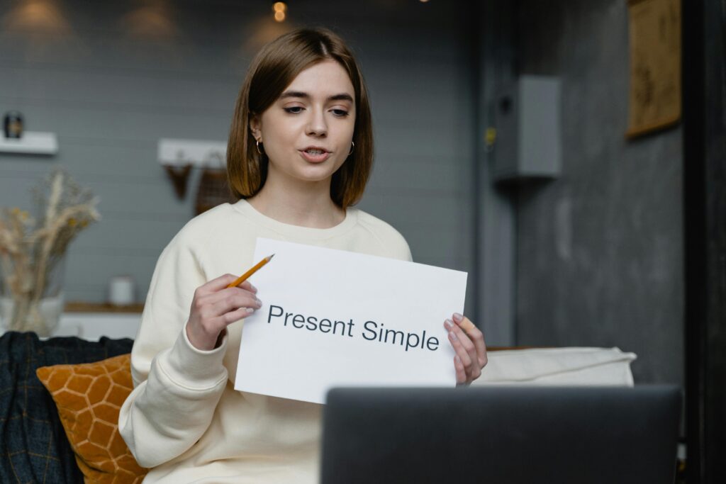 pexels-photo-6671683-6671683 Young woman teaches English grammar online, holding a 'Present Simple' note and pencil.