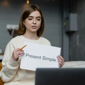 Young woman teaches English grammar online, holding a 'Present Simple' note and pencil.