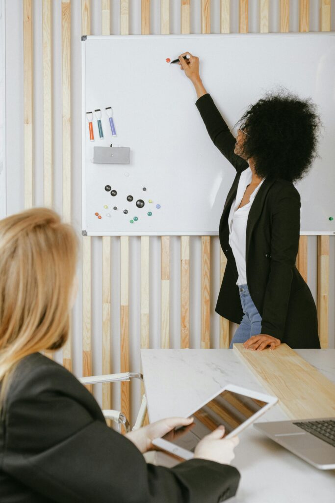 pexels-photo-3727512-3727512 Two women engaged in a business presentation with a whiteboard in a modern office setting.