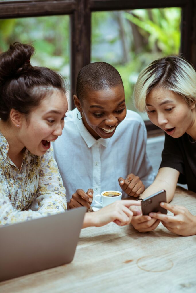pexels-photo-4353618-4353618 Amazed surprized multiethnic female friends in casual clothes with cup of coffee browsing smartphone while sitting at wooden table with laptop against window