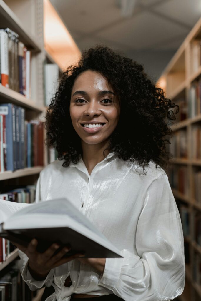 Smiling African American woman holding a book in the library, embodying education and learning.